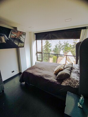A bedroom with a bed, a TV mounted on the wall, a window with a view of buildings and trees, and a small table with a statue. - Beautiful and Spacious Apartment in Chapinero Bogota DC (Bogotá, D.C.)