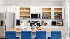 Une cuisine moderne avec des meubles blancs, un réfrigérateur en acier inoxydable, un four à micro-ondes, une cuisinière et un grille-pain. Des chaises bleues sont disposées autour d’un îlot central sur lequel se trouve un saladier.