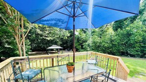 A glass-top table with chairs, a blue umbrella, and a wooden deck with a gazebo in the background.