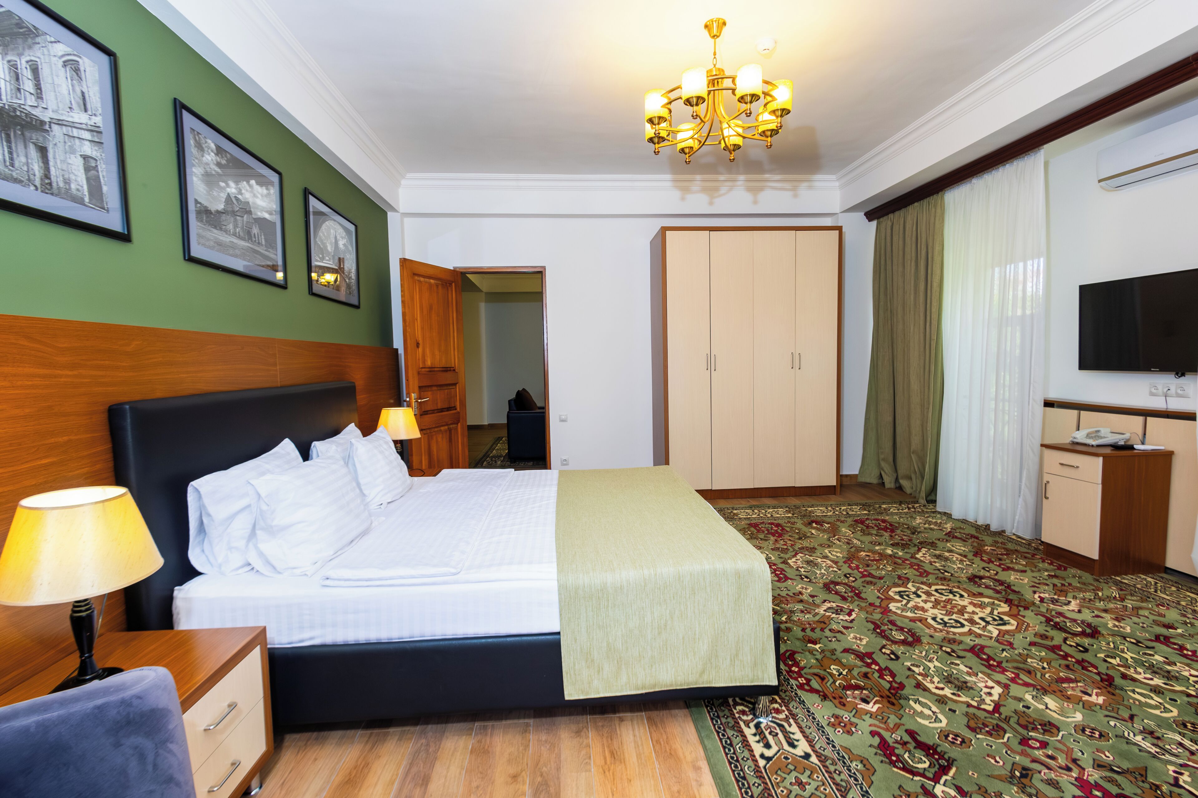 A hotel room with a bed, a desk with a lamp, a television, a wardrobe, and a carpeted floor.