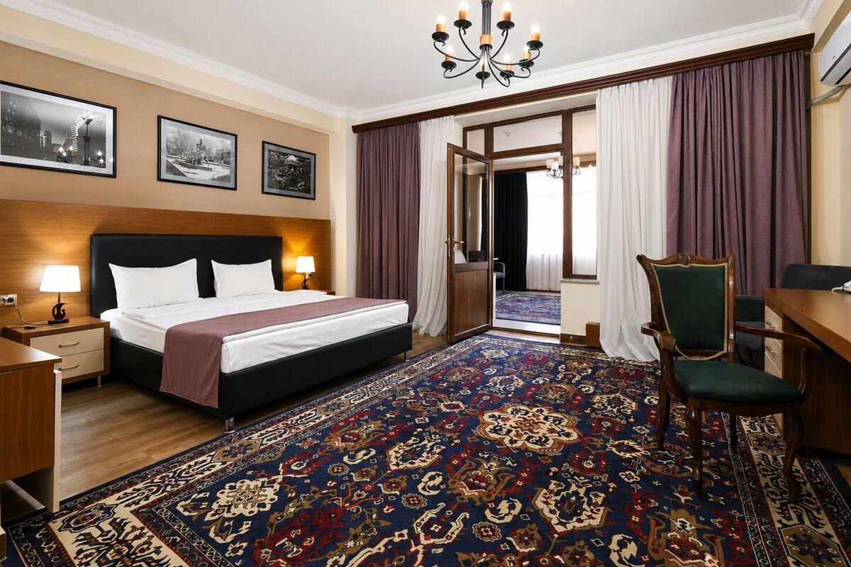 Habitación de hotel con cama, escritorio, silla, candelabro y alfombra grande con estampado.