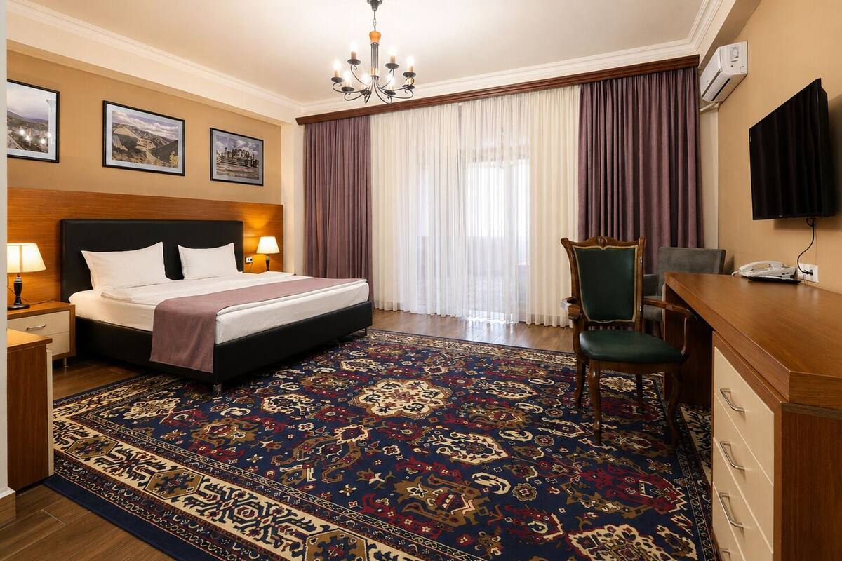 Habitación de hotel con cama, escritorio, silla, candelabro, televisor y un amplio ventanal con cortinas.