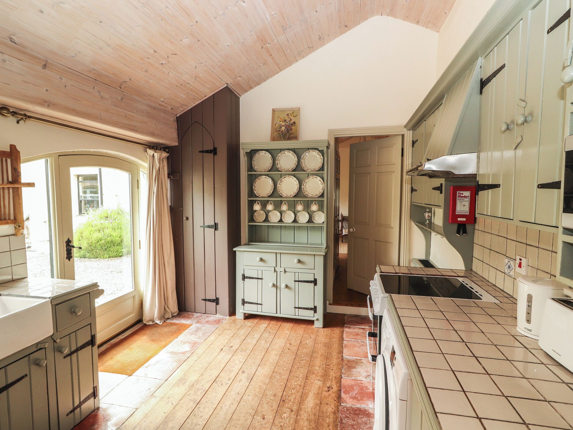 A kitchen with wooden flooring, a white stove, a cabinet with plates, and a door leading outside.