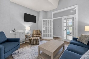 Un salon moderne avec un téléviseur à écran plat, deux canapés, une table basse, un fauteuil à motifs, une table d’appoint et une porte donnant sur un balcon.