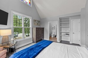 Une chambre à coucher avec un lit, une commode, un téléviseur, une fenêtre et une salle de bain visible à travers une porte ouverte.