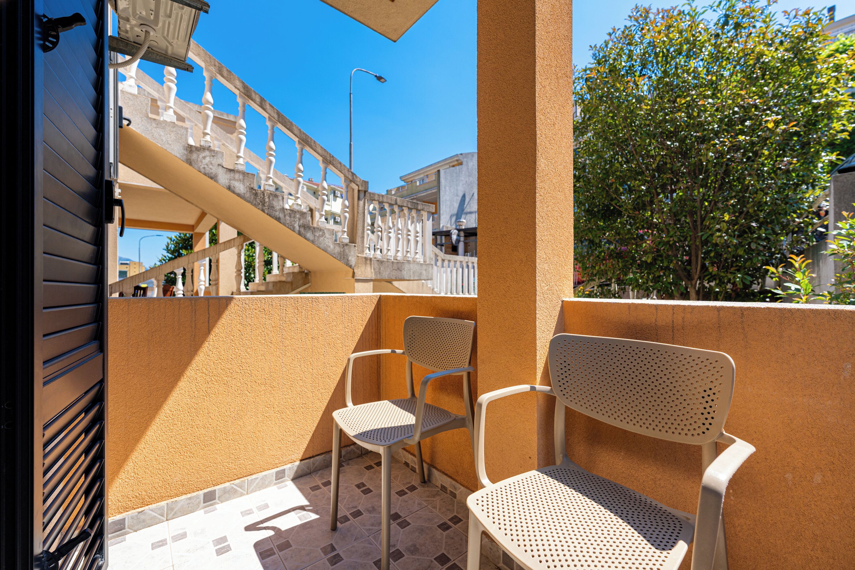 A balcony with two chairs and a table, a staircase, and a view of a tree and buildings.
