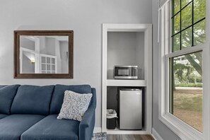 Ein kompakter Wohnraum mit einer Couch, einer Mikrowelle und einem Kühlschrank auf einer eingebauten Ablage, einem Spiegel an der Wand und einem Fenster mit Blick auf Grünflächen.