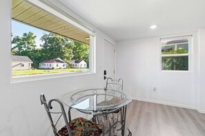 A room with a glass table and metal chairs, a window with a view of houses, and a door.