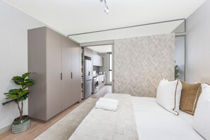 Een moderne slaapkamer met een bed, een kamerplant, een kledingkast en uitzicht op een keuken.