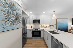 A modern kitchen with stainless steel appliances, a white countertop, and a large decorative painting. - Golden Aura | Pool Front | Gulf View | Sleeps 6 (Miramar Beach)