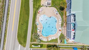 Eine Vogelperspektive zeigt ein Hotel mit einem zentral gelegenen Schwimmbad, einem Parkplatz und einer angrenzenden Straße.