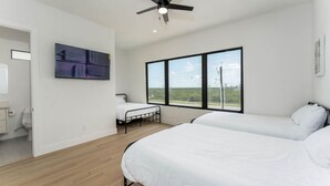 Une chambre moderne avec un lit, une petite table, un ventilateur de plafond, une fenêtre donnant sur un beau paysage et un tableau accroché au mur.