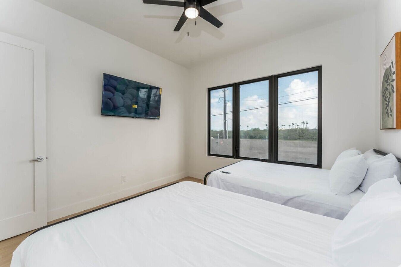 Quarto com duas camas, um ventilador de teto, uma janela grande com vista e um quadro na parede.