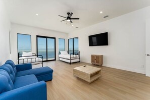 A modern living room with a blue sofa, a small wooden coffee table, a white chair, a TV mounted on the wall, and a balcony with a view of the sea. - 4,000 SF Waterfront Home w/ Pool, Sleeps 22 (Port Isabel)