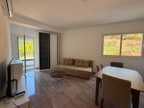 Ein Zimmer mit einer Couch, einem Esstisch, einem Fernseher und einer Schiebetür, die auf einen Balkon führt.