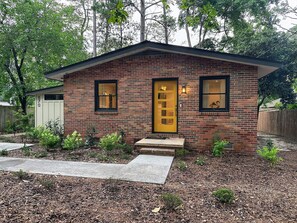 Une petite maison en briques avec une porte jaune, deux fenêtres et un jardin devant la maison, avec du paillis et de petites plantes.