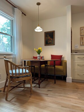 Une pièce avec un plancher en bois, une table à manger avec des chaises, un canapé, une petite table sur laquelle est posé un vase de fleurs, un luminaire suspendu et un tableau accroché au mur.