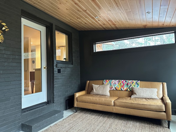 Ein modernes Interieur mit dunkler Wand, Holzboden und einem beigefarbenen Ledersofa mit bunten Kissen, sowie ein Fenster mit Blick.