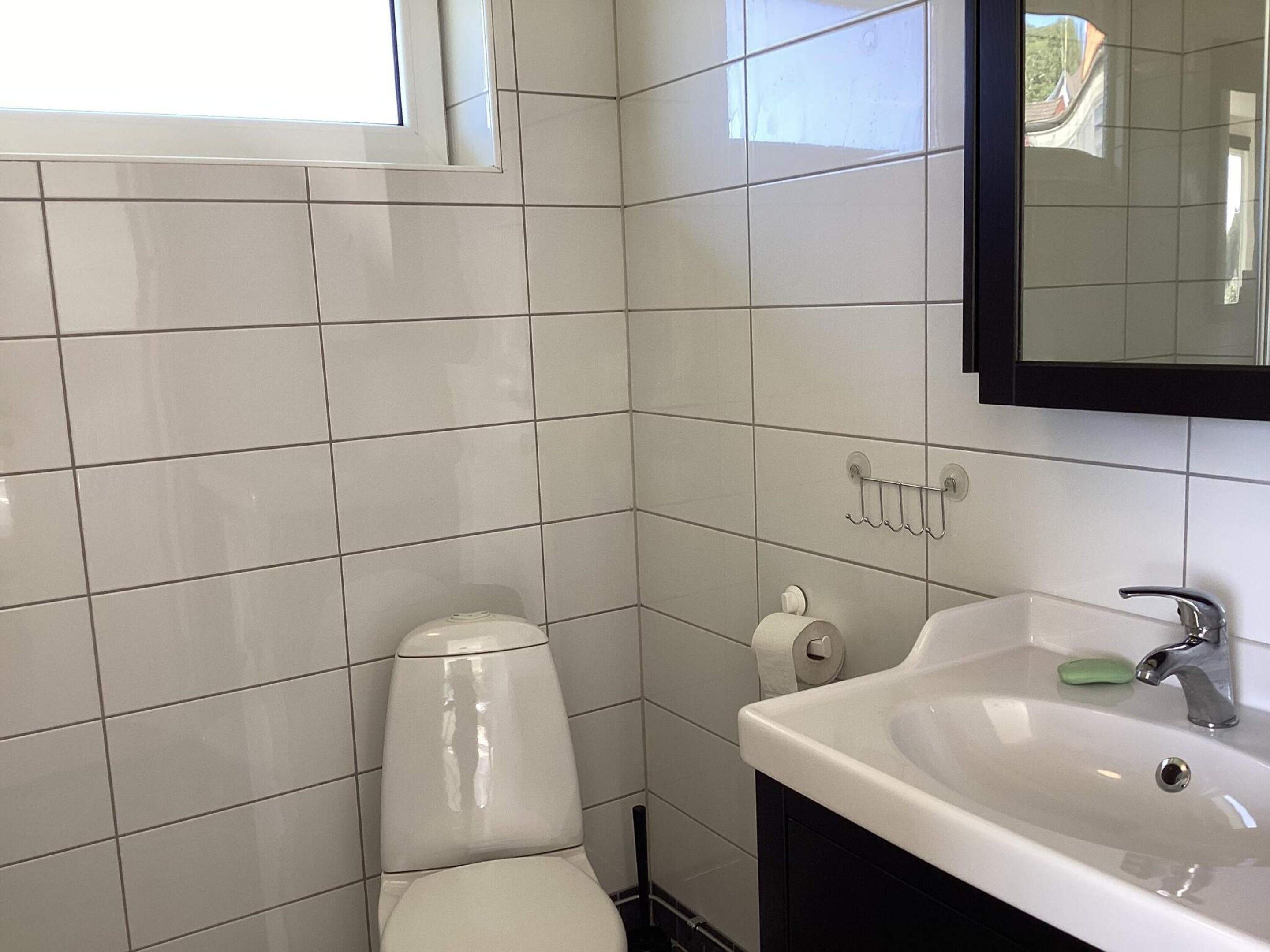 Un bagno con piastrelle bianche, un water, un lavandino con un porta sapone e uno specchio sopra il lavandino.
