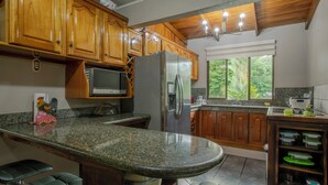 A kitchen with wooden cabinets, a granite countertop, a refrigerator, a microwave, and a window with a view of trees. - Gato&Culla House (La Fortuna)