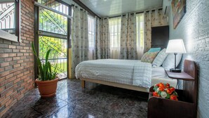 A bedroom with a bed, a nightstand, a lamp, a potted plant, and a window with curtains. - Gato&Culla House (La Fortuna)