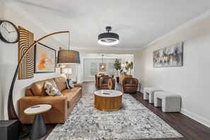 Ein modernes Wohnzimmer mit einer Couch, einem Couchtisch, Hocker und einem Essbereich im Hintergrund.