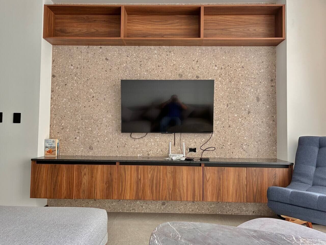 A modern living room with a flat-screen TV mounted on a textured wall, wooden cabinets below, and a blue sofa.