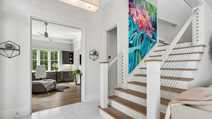 Un interior moderno con una escalera, un cuadro abstracto colorido y vista hacia una sala de estar con sofá y planta.