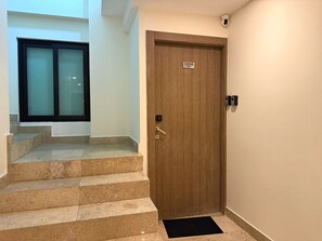Una puerta de madera con una placa de número, un felpudo negro al pie de una escalera y un ventanal con vidrio esmerilado.