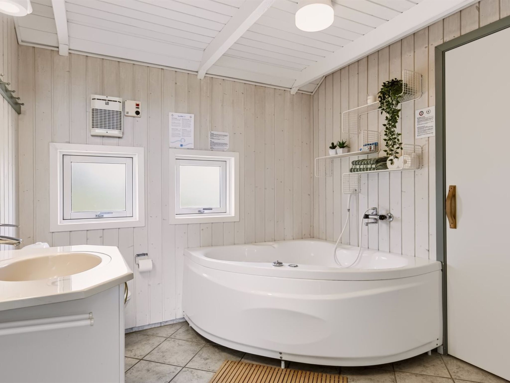 A bathroom with a large bathtub, a sink, and a shower. There are two windows, a wall-mounted air conditioner, and a shelf with plants.