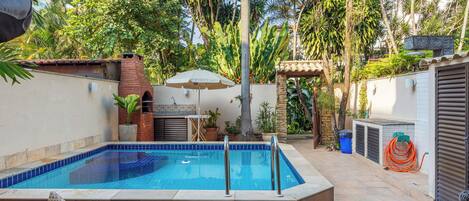 A swimming pool with a tiled deck, surrounded by a garden with palm trees and a brick structure.