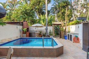 Ein Schwimmbecken mit gefliestem Deck, umgeben von einem Garten mit Palmen und einem Mauerwerk.