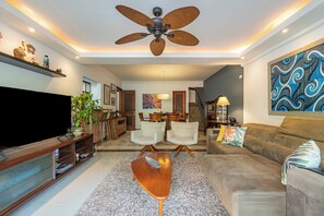 Ein modernes Wohnzimmer mit Flachbildfernseher, Deckenventilator, Sofa, Couchtisch und einem Essbereich mit Stühlen und Tisch.