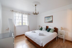 A bedroom with a wooden bed, white linens, a chandelier, a painting, and a bedside table with a lamp. - Holiday Home for 9 in St. Renan (Saint-Renan)