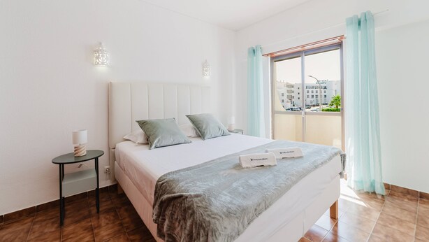 A hotel room with a bed, a nightstand, a lamp, a window with curtains, and a view of buildings outside. - Blue Stay - 1º D by HelloVacations (Albufeira)