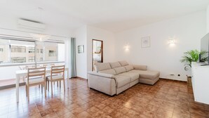 A modern living room with a beige sectional sofa, a dining table with chairs, a large window, and a potted plant. - Blue Stay - 1º D by HelloVacations (Albufeira)
