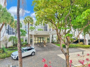A hotel entrance with a paved driveway, palm trees, and a white car parked in front. - Top-Floor Near Beach Condo, Pool + Skyline Views (Sunny Isles Beach)