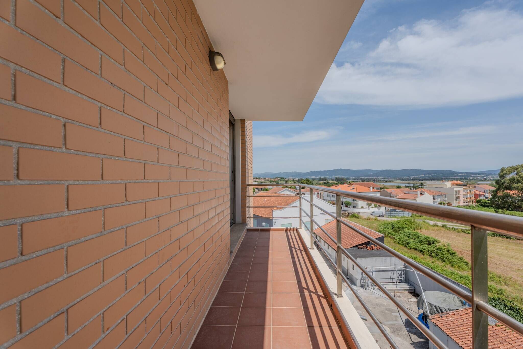 A balcony with a brick wall, metal railing, and a clear view of buildings and hills in the distance.