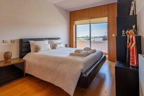 Ein modernes Hotelzimmer mit einem großen Bett, einem Nachttisch, einer Kommode und Blick auf die Stadt durch große Fenster.