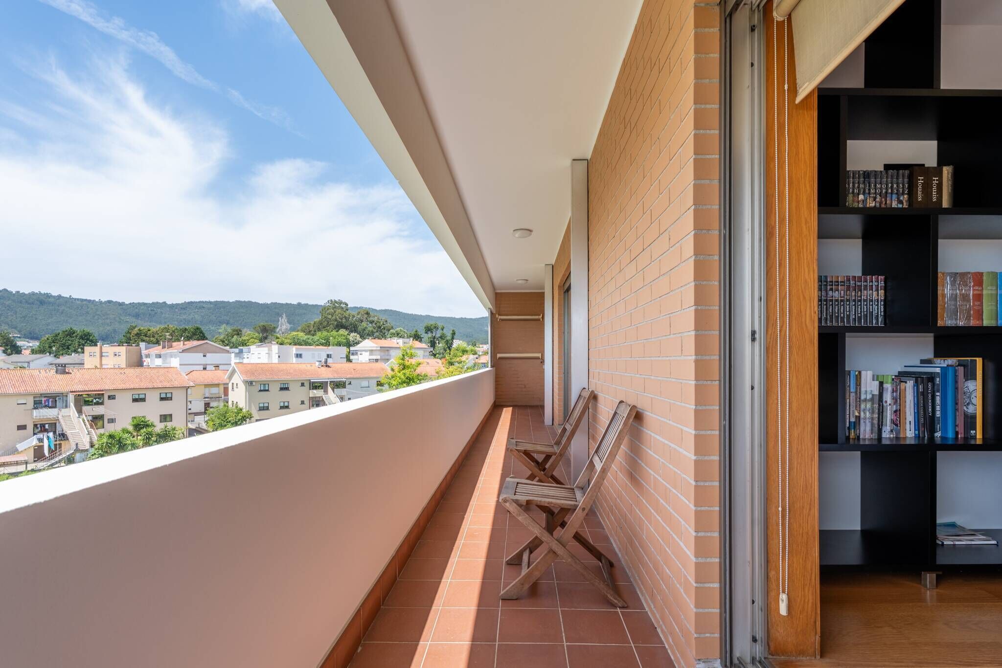 Ein Balkon mit Blick auf Gebäude und Hügel, ein Bücherregal sowie zwei Holzstühle.