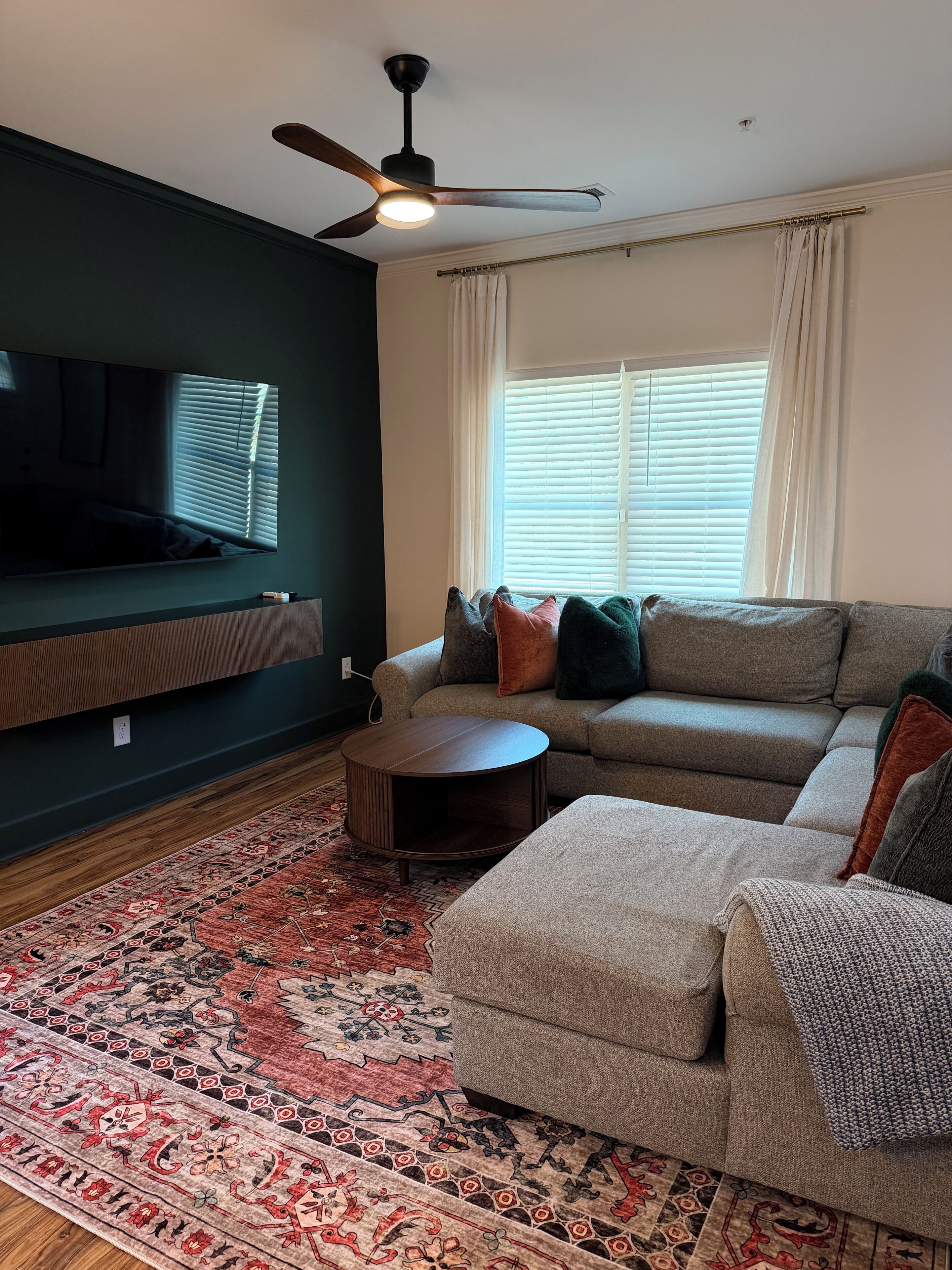 A living room with a sofa, ottoman, TV, ceiling fan, and patterned rug.