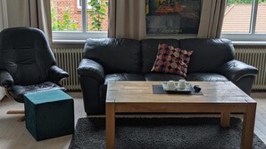 Uma sala de estar com um sofá de couro preto, uma mesa de centro de madeira com um objeto decorativo, um banquinho preto e uma janela com vista para um edifício de tijolos.