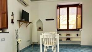 A room with a white chair, a small table, a TV on the wall, a window with wooden shutters, and a tiled floor. - Casa Tasca, a cozy and quiet apartment with a large terrace. (Ravello)