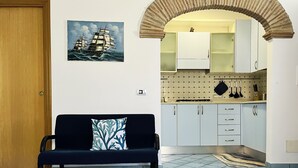 A room with a sofa, a painting of ships, a kitchen with cabinets, and a tiled floor. - Casa Tasca, a cozy and quiet apartment with a large terrace. (Ravello)
