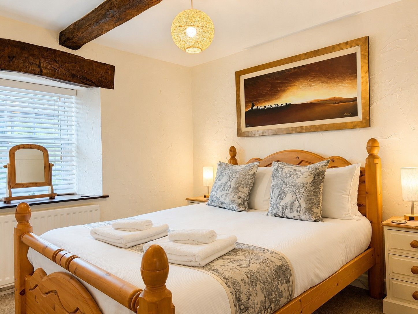 A bedroom with a wooden bed, a nightstand, a bedside lamp, a window with blinds, and a painting on the wall.