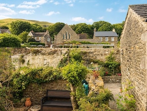 A stone building with a wooden bench, a small door, and a garden area with plants and a hose. - Sheepfold Cottage – Cosy Countryside Escape for up to 4 Guests (Sedbergh)