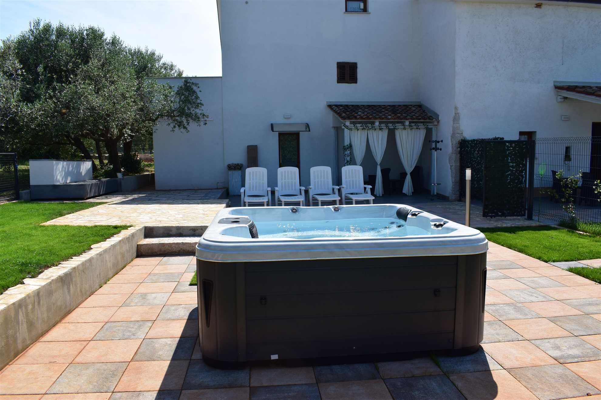 Un jacuzzi dans un jardin, avec une terrasse carrelée, des chaises blanches et un bâtiment en arrière-plan.