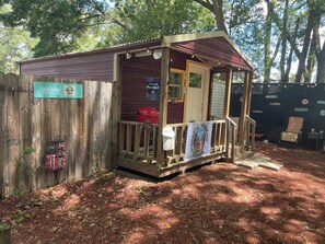 Une cabane en bois rustique, dotée d’une véranda, d’une pancarte d’accueil et d’un panneau indiquant la présence d’un bar et d’un coin barbecue dans le jardin.