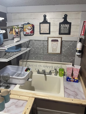 A kitchen with a sink, a wine glass, a dish rack, a framed coffee cup, and popcorn boxes. - Charming Rustic Tiny Home – Private 1 Ac Oasis - Yulee, FL (Yulee)