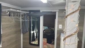 Een kamer met een houten vloer, een kledingrek, een spiegel, een klein krukje en een deur naar een andere kamer.
